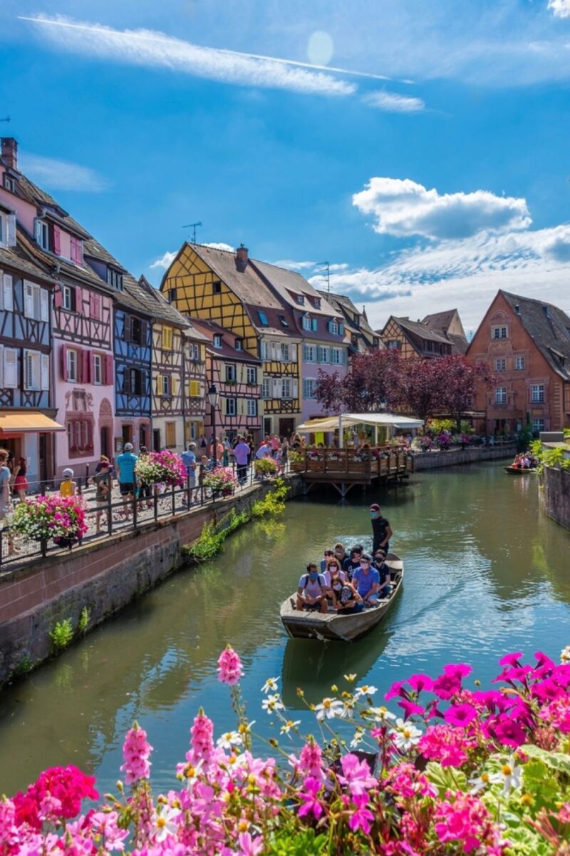 A vibrant scene in a European town with traditional half-timbered houses in a variety of colors lining a canal. Tourists enjoy a leisurely boat ride on the calm water, surrounded by lush pink and white flowers under a clear blue sky with wispy clouds.