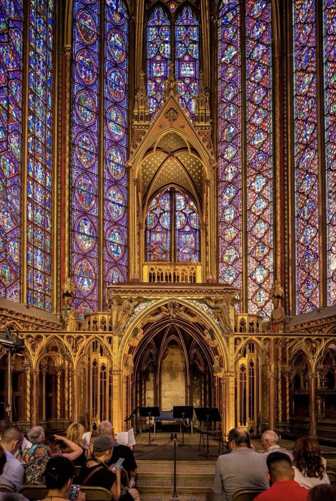 Visitors seated inside the Sainte-Chapelle in Paris, awaiting a concert, surrounded by the chapel's stunning gothic architecture and radiant stained glass windows that cast colorful patterns across the ornate interior.