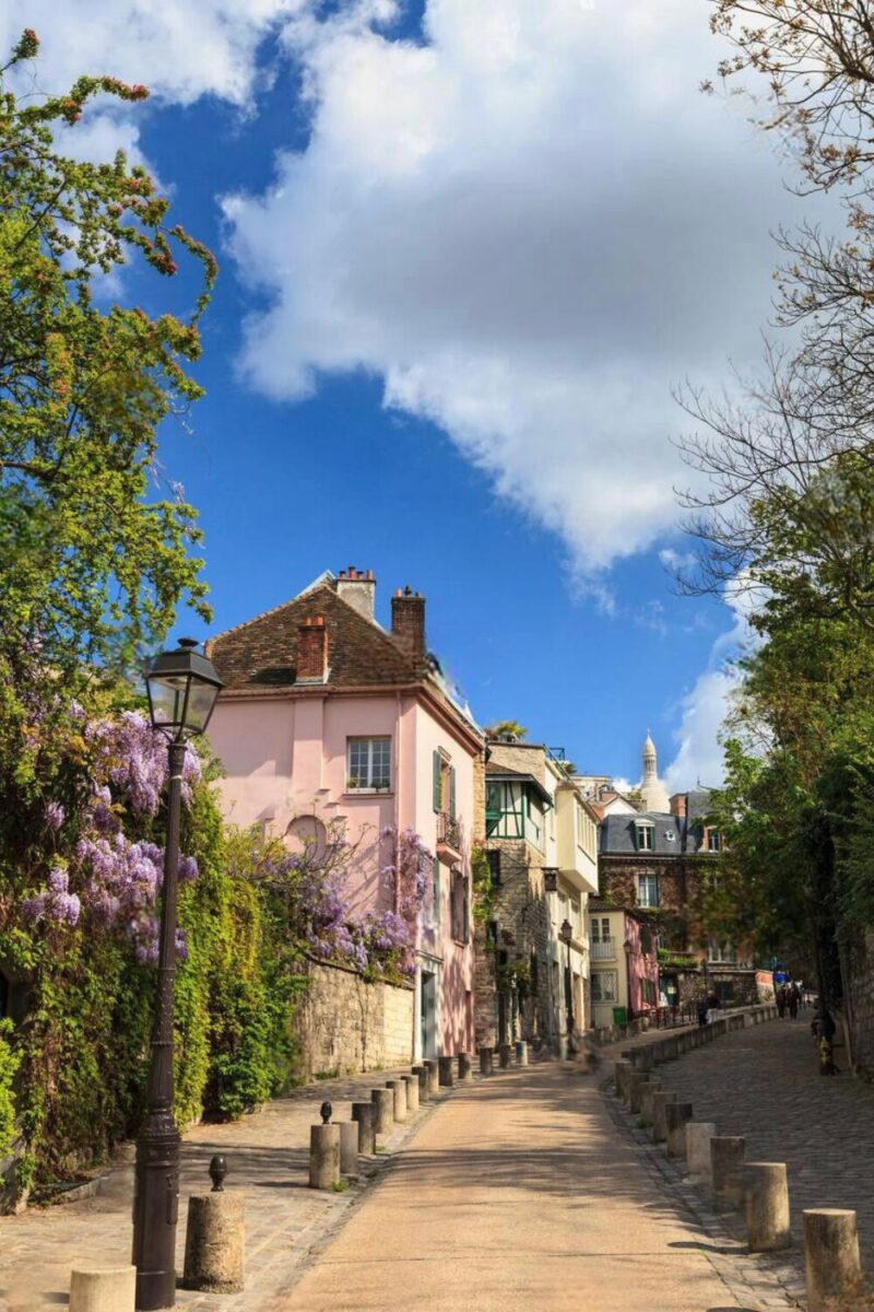 The photograph presents a serene view of Rue de l'Abreuvoir in Montmartre, Paris. The street is lined with charming pastel-colored buildings and blooming wisteria. Traditional Parisian lampposts border the cobblestone pathway, leading towards a distant spire. The composition is framed by a bright blue sky dotted with fluffy clouds, adding to the idyllic and peaceful ambiance of this picturesque neighborhood.