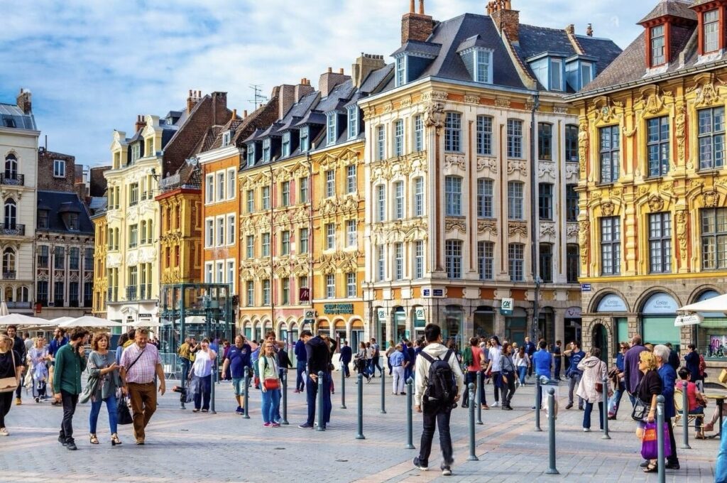 Things to Do in Lille: Vibrant city square bustling with people, flanked by historic buildings with ornate facades in a mix of baroque and classical architecture, under a clear blue sky.