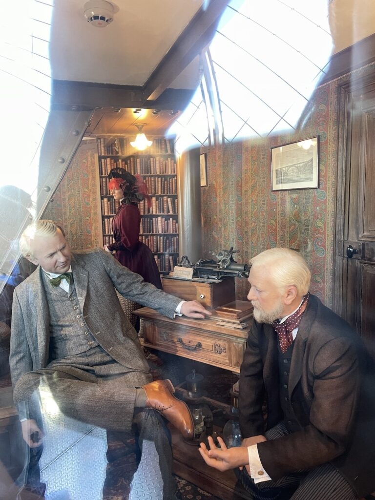 A look inside Gustave Eiffel’s secret apartment at the top of the Eiffel Tower, featuring wax figures of Eiffel and Thomas Edison in a historical setting with vintage furniture, patterned wallpaper, and a bookshelf filled with classic books.