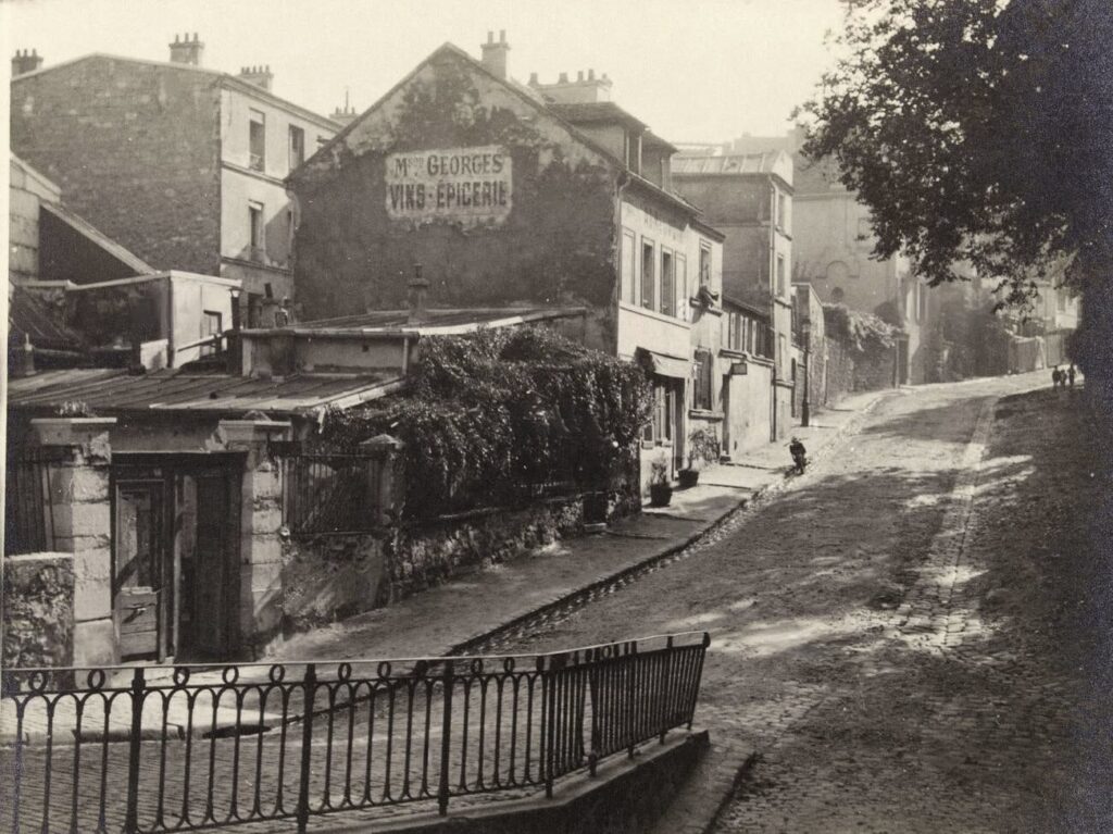 This is a vintage black and white photograph depicting a quiet, cobblestone street with old buildings and a prominent advertisement on a wall for "Maison Georges Vins-Épicerie". A lone figure can be seen in the distance, and there's a tree casting a shadow on the right. The image has an old-world charm, with ivy creeping over walls and a rustic iron railing in the foreground.