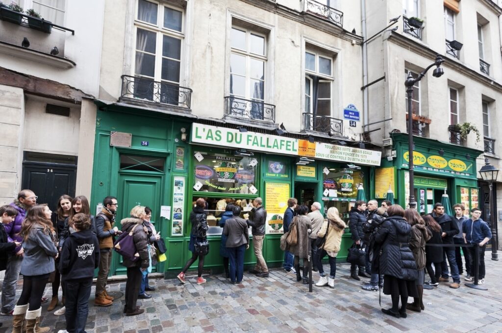 24 Hours in Paris: A bustling scene in front of L'As du Fallafel, a famous falafel restaurant in Paris, with a crowd of people queuing outside the vibrant green storefront, located in the historical Marais district.