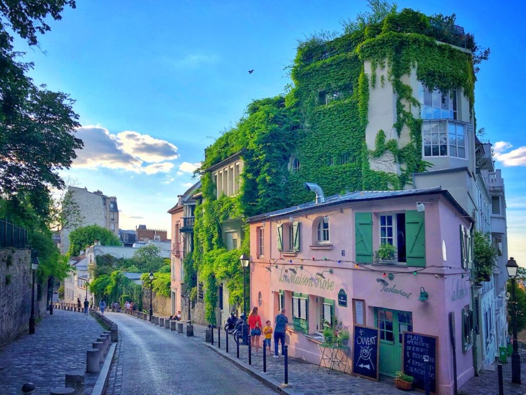 This image captures the charming and picturesque Rue de l'Abreuvoir in Montmartre, Paris, with the famous La Maison Rose restaurant at the corner. The cobblestone street gently curves, lined with lush green ivy-covered buildings under a serene blue sky. Pedestrians can be seen walking along the sidewalk, enjoying the quaint ambiance of this iconic neighborhood.