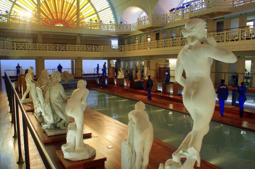 Spacious gallery featuring an array of classical statues displayed around a reflective pool, with visitors admiring the art under a striking stained glass ceiling.