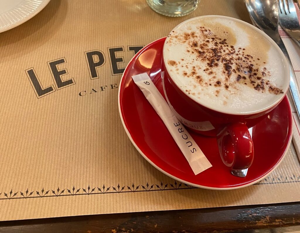 A cappuccino with frothy milk and sprinkled chocolate rests in a vibrant red cup on a saucer, accompanied by a sugar packet on a table with 'LE PETIT CLER CAFÉ' printed on the placemat, evoking the cozy ambiance of a Parisian café.