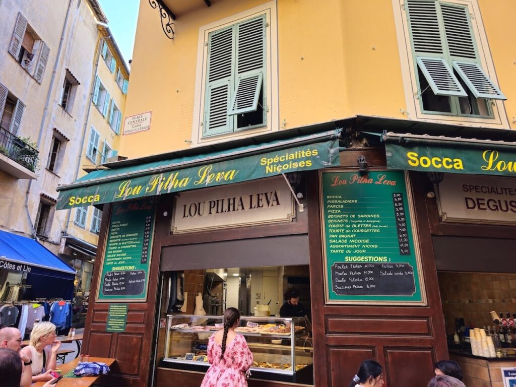 The corner view of Lou Pilha Leva, a local eatery in Nice, France, known for its 'Spécialités Niçoises' including socca, as indicated by the prominent green signs. Patrons are seated outside the yellow building with traditional green shutters, enjoying the Niçoise culinary experience.