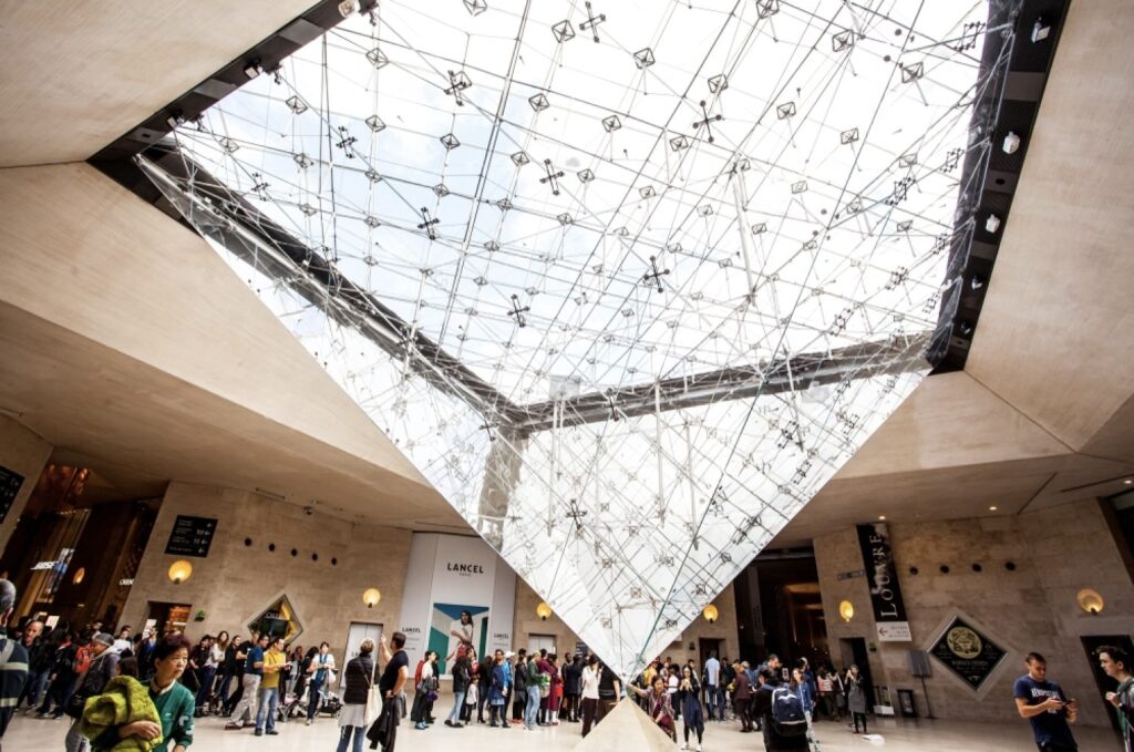 24 Hours in Paris: Visitors gather beneath the inverted glass pyramid at the Louvre Museum, with its intricate network of steel and glass casting geometric patterns above the bustling lobby area.