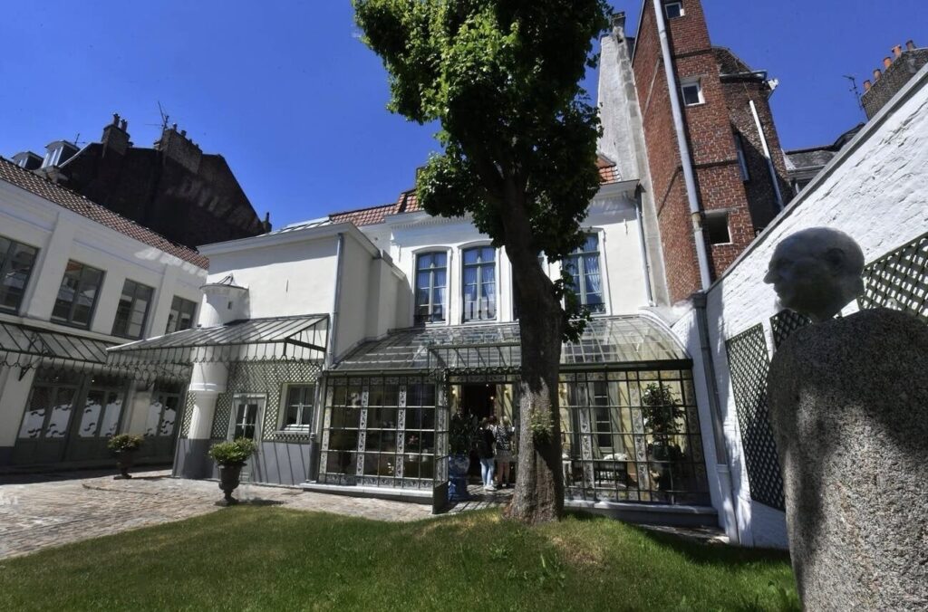 Things to Do in Lille: Luxurious seaside villa with a columned balcony overlooking a calm bay, accompanied by a mature pine tree and a solitary white chair set on a gravel path.