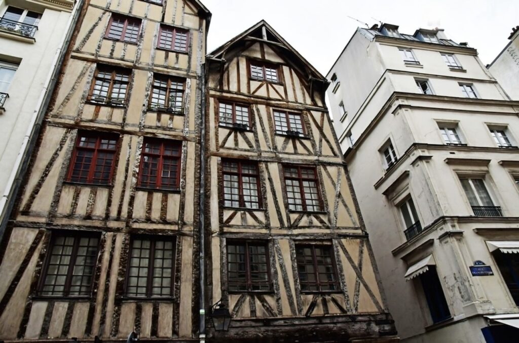 Historical half-timbered houses, Maison du Faucheur and Maison du Mouton, in Paris, with their distinctive wooden beam structures and patterned exteriors. The aged facades feature a combination of closed and open shutters on windows with red and white panes, set against the backdrop of more modern buildings.