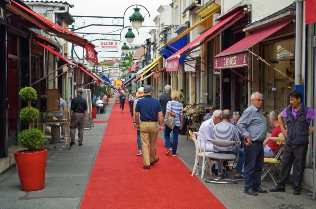 Bustling activity in a traditional Parisian market street, lined with colorful shop awnings and a red carpet, as people browse and socialize, capturing the charm of local French commerce.