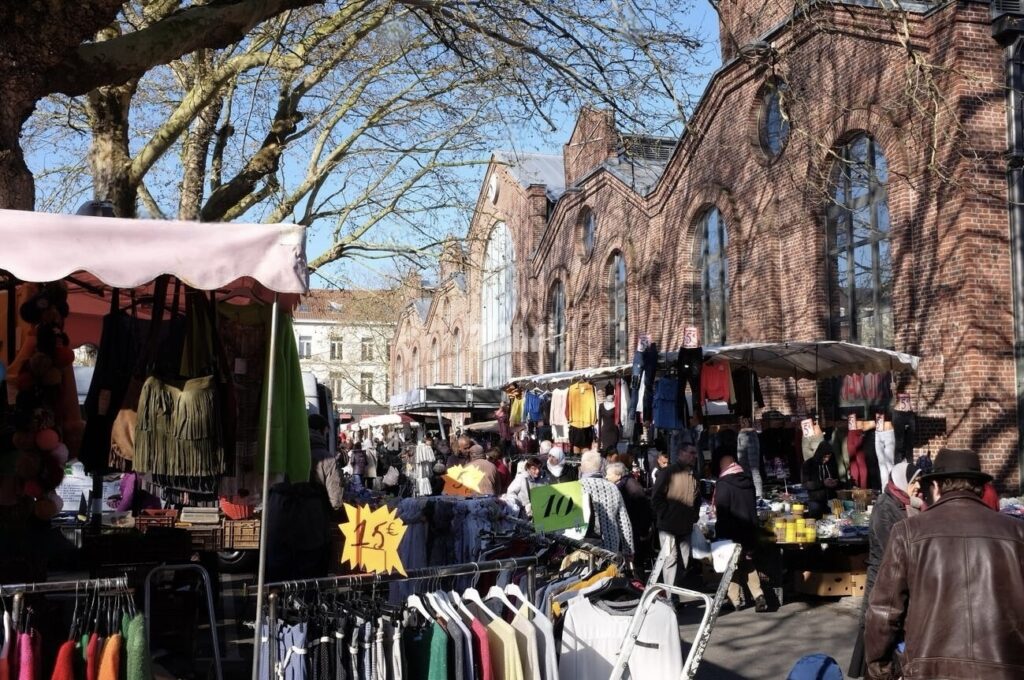 Outdoor market scene with a variety of clothing and goods on display under makeshift stalls, set against the backdrop of a historic brick building, with shoppers milling about in a tree-lined street.
