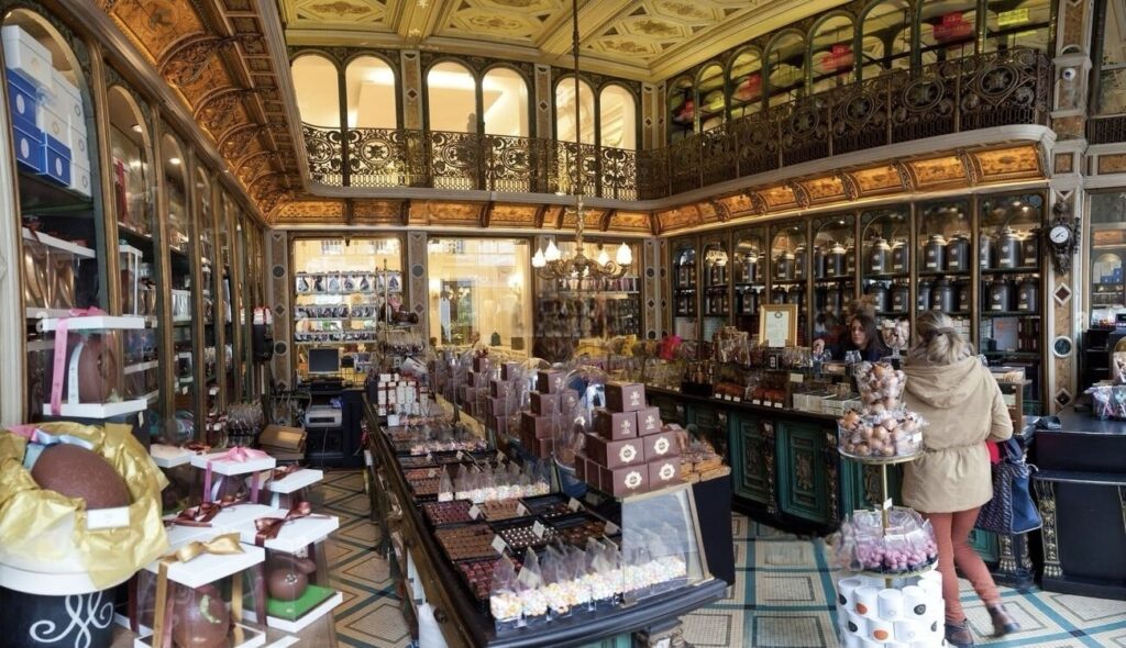 Things to Do in Lille: Elegant interior of a traditional chocolate shop with ornate gold ceiling and wood-paneled walls, showcasing a variety of chocolates and confectionaries on display counters, with customers browsing the selection.