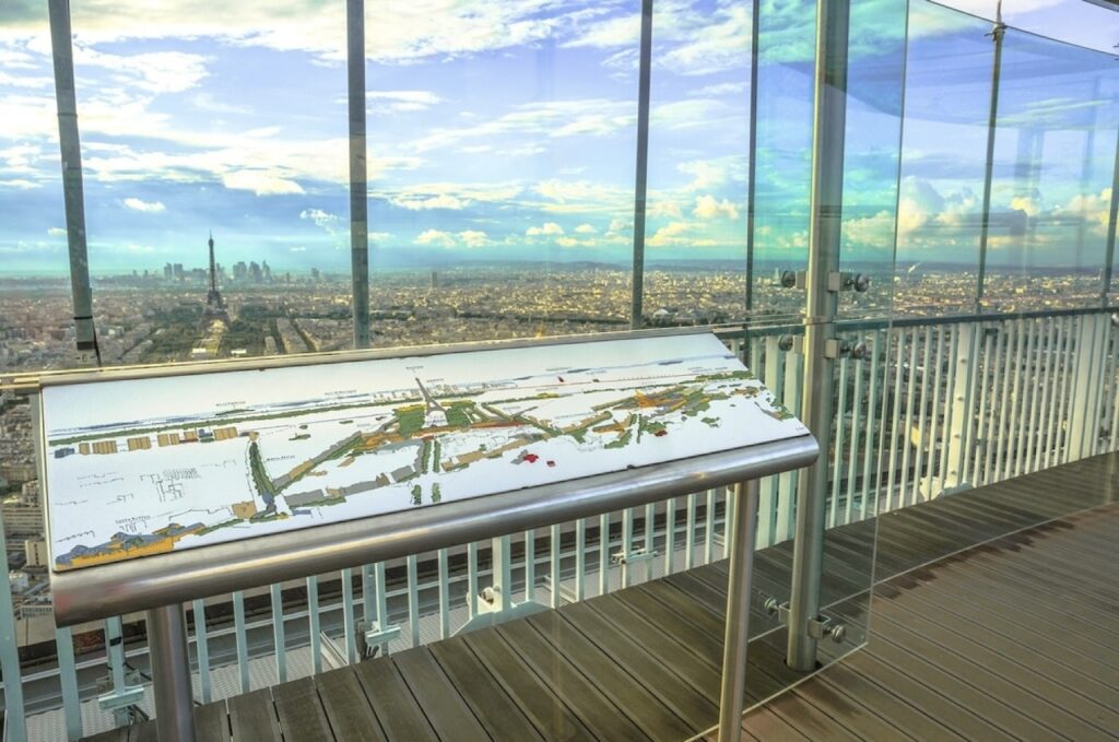 hidden gems in paris: Panoramic view of Paris from the Montparnasse Tower viewing deck, with a detailed map in the foreground identifying key landmarks. The Eiffel Tower stands out against the cityscape under a cloudy sky, seen through the clear glass barriers of the observation deck.
