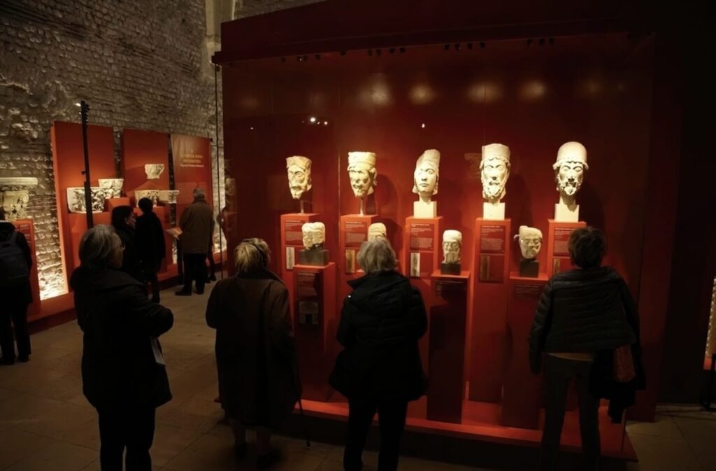 Visitors observing ancient sculpted heads displayed in red-lit cases at the Musée de Cluny in Paris, set against the rustic stone walls of the medieval building. The exhibit showcases a collection of historical artifacts, creating a contemplative atmosphere.