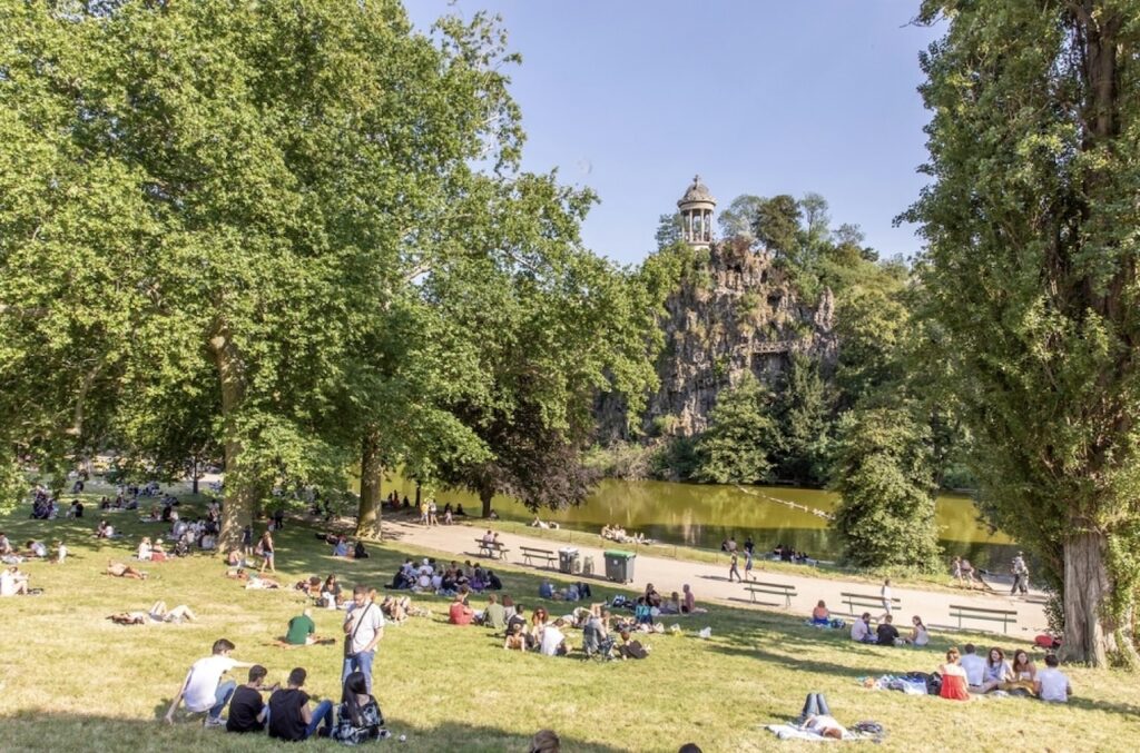 A vibrant day at Parc des Buttes-Chaumont in Paris, with people scattered across the grassy slopes enjoying picnics and relaxation, overlooking the serene lake and the towering Temple de la Sibylle on a cliff.