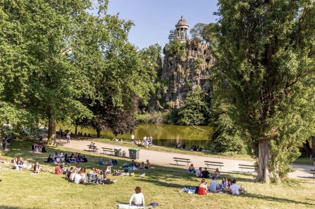 A sunny day at Parc des Buttes-Chaumont in Paris, with people lounging and socializing on the grassy slopes. In the background, the park's distinctive rocky cliff and the Temple de la Sibylle perched atop it overlook a serene lake.