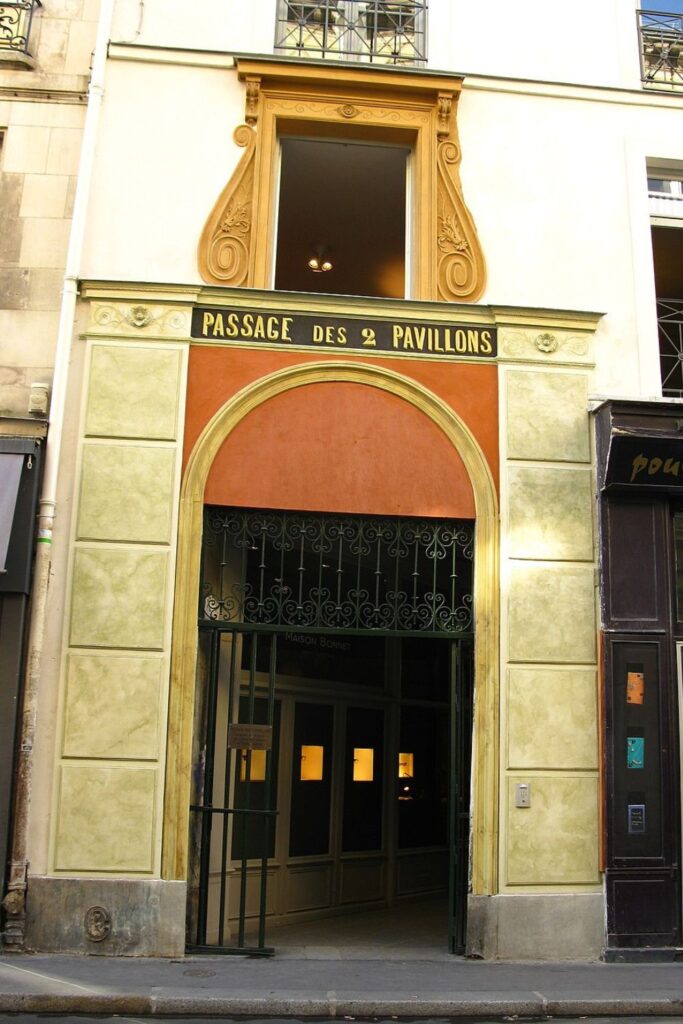 The ornate entrance to Passage des Deux Pavillons in Paris, with 'PASSAGE DES 2 PAVILLONS' lettered above a wrought iron gate. The classical architectural details and muted color palette convey the historical elegance of this Parisian landmark. A glimpse into the passage reveals a warm, inviting interior with visible lights.