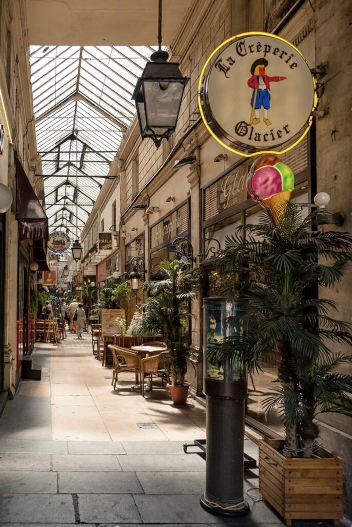 Quaint corner of the Passage des Panoramas in Paris, showcasing a creperie with a bright, illustrated sign. A classic Parisian street lamp and vibrant green plants enhance the cozy, inviting atmosphere. The passage's glass roof and architectural details form a picturesque backdrop to this serene urban oasis.