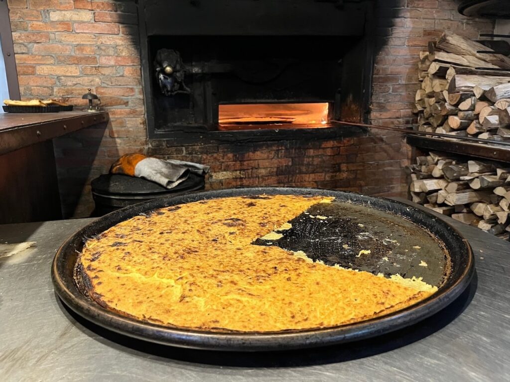 A large, half-eaten socca, which is a traditional chickpea flour pancake, on a round black baking tray, with a wood-fired oven and stacked firewood in the background, indicating a rustic setting likely in a restaurant.