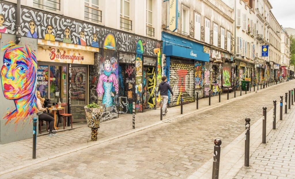hidden gems in paris: Vibrant street art adorns the storefronts of Rue Dénoyez in Paris, known for its dynamic graffiti and murals. A pedestrian strolls past the colorful urban canvas, while a patron enjoys a drink outside the eclectic 'Le Barbouquin' café.