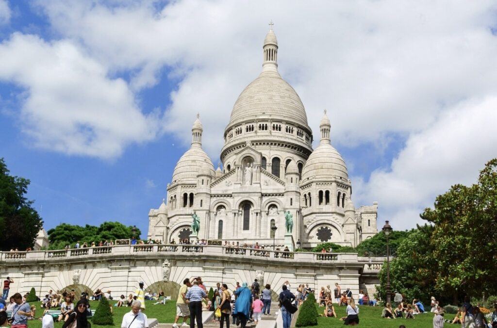 The photo showcases the Basilica of the Sacré-Cœur, perched on the Montmartre hill in Paris. The majestic white domes of the basilica rise against a bright blue sky with fluffy clouds. In the foreground, the terraced lawn is filled with tourists and locals enjoying a sunny day, some sitting and others walking, with greenery framing the scene.