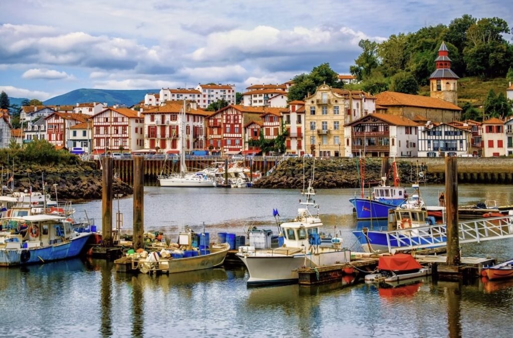 The serene port of Saint-Jean-de-Luz, France, with a flotilla of fishing boats moored in calm waters, and the traditional Basque architecture providing a colorful backdrop under a partly cloudy sky.