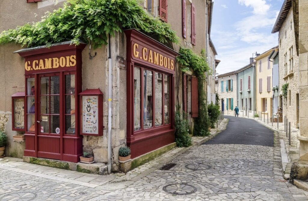 A quaint corner of a French village with a shop named 'G. CAMBOIS' featuring red storefronts and windows, adorned with greenery. The cobblestone street leads past colorful houses and a solitary figure walking away, enhancing the serene, old-world charm of the scene.