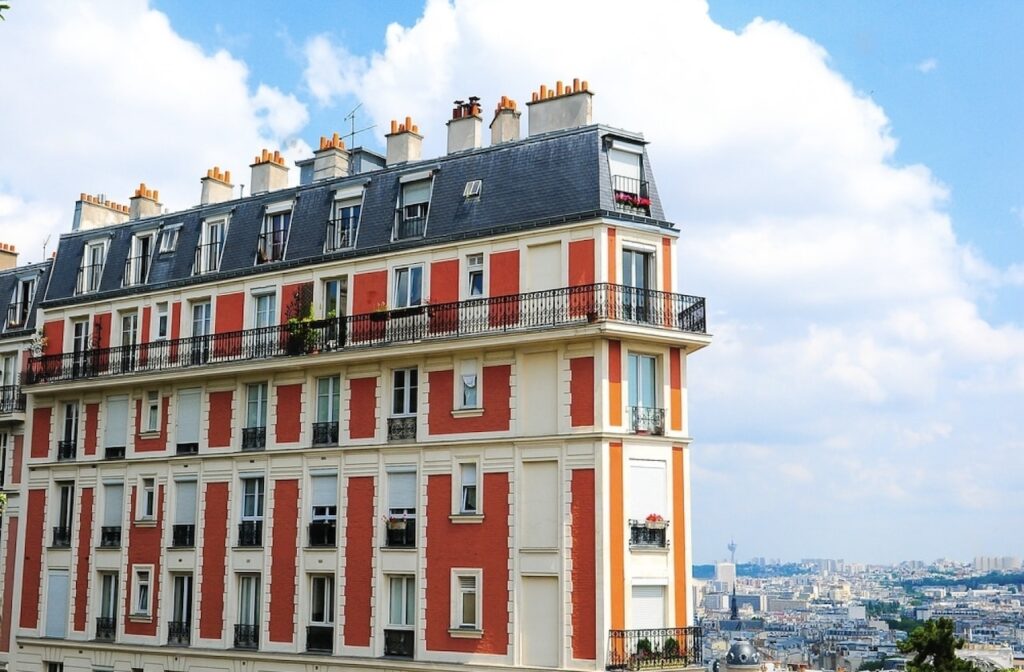 A classic Parisian building with vibrant red and cream facade, blue slate roof, and ornate wrought-iron balconies, set against a backdrop of the Paris skyline under a partly cloudy sky.