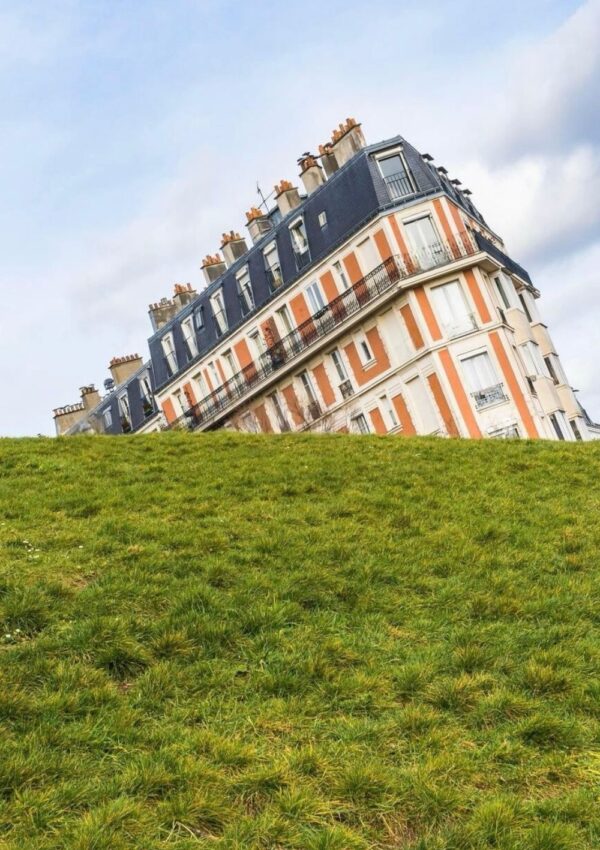 An unusual perspective of a traditional Parisian building creating the optical illusion of a sinking house, with its base obscured by a grassy hill, set against a blue sky with light clouds.