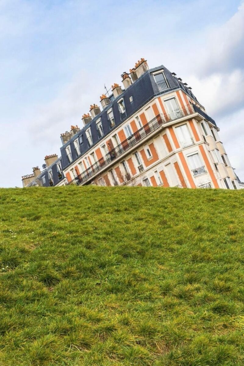 An unusual perspective of a traditional Parisian building creating the optical illusion of a sinking house, with its base obscured by a grassy hill, set against a blue sky with light clouds.