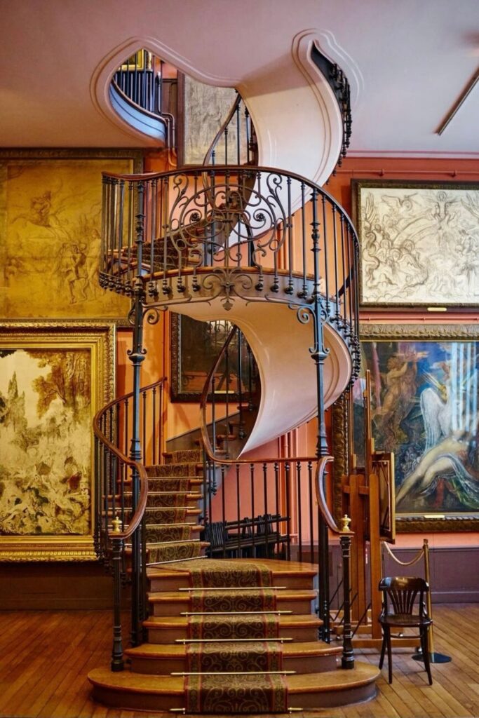 An ornate spiral staircase with intricate iron balusters ascends within a warmly lit museum room adorned with large, framed artwork depicting classical scenes. The elegant wooden steps are carpeted with a patterned runner, and a solitary wooden chair sits at the base of the stairs, contributing to the room's historical ambiance.