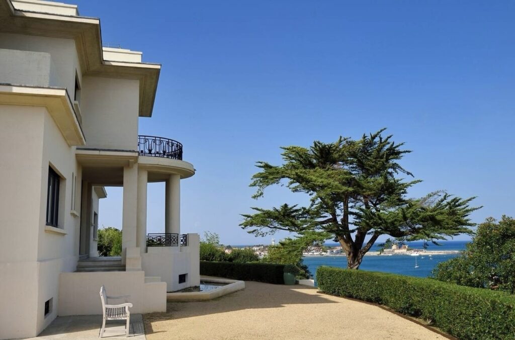 Elegant white villa with a circular balcony overlooking a serene seascape, flanked by a mature pine tree, with a clear blue sky above and a quaint coastal town in the distance.