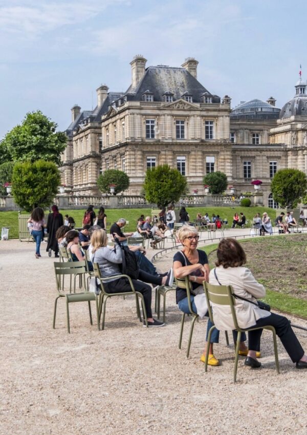 Tourists and locals enjoy a leisurely time in Luxembourg Garden, Paris, seated on the iconic green metal chairs against the backdrop of the grand Senate building, under the verdant trees on a bright day.