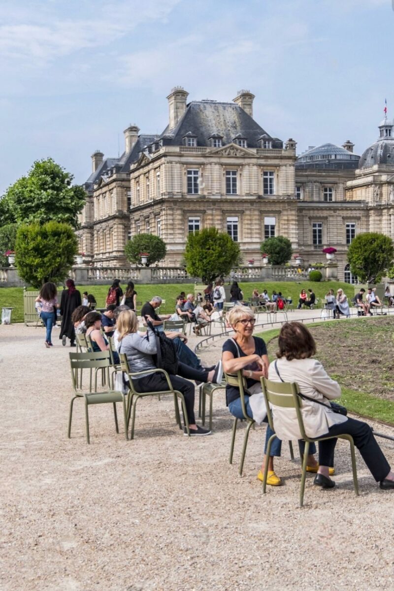 Tourists and locals enjoy a leisurely time in Luxembourg Garden, Paris, seated on the iconic green metal chairs against the backdrop of the grand Senate building, under the verdant trees on a bright day.