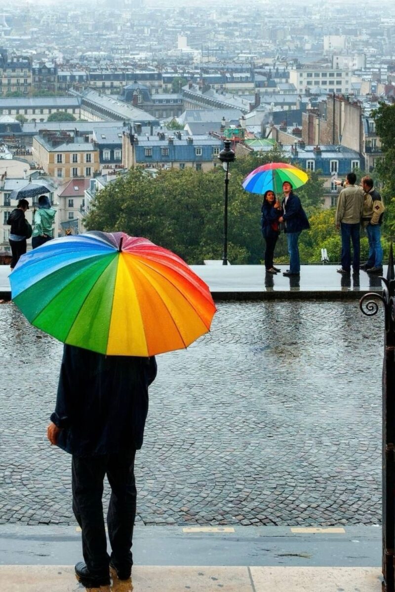 A person holding a vibrant rainbow-colored umbrella overlooks the misty Paris cityscape from a high vantage point, with other individuals holding umbrellas and enjoying the view on a rainy day.