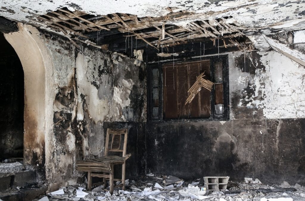 Interior of an abandoned building showing extensive fire damage. Charred walls and a collapsed ceiling reveal the wooden beams, with a solitary damaged wooden chair and debris scattered across the floor.