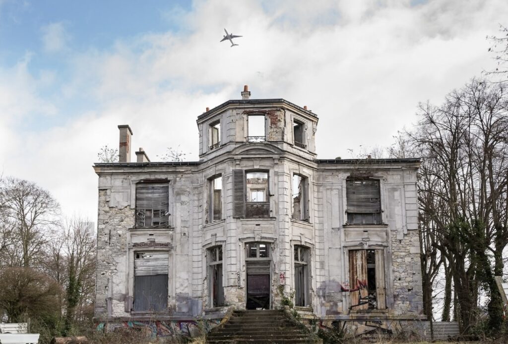 Facade of an abandoned, dilapidated two-story mansion with overgrown vegetation and graffiti, under a cloudy sky with a jet airplane flying overhead.