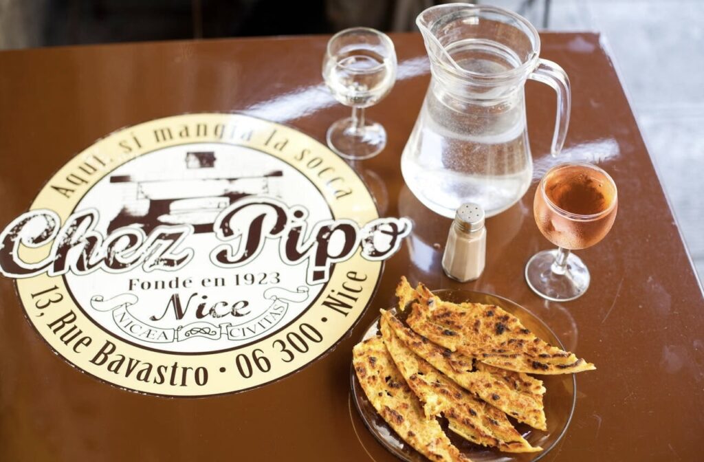 A table at Chez Pipo in Nice displays their famous socca, a golden chickpea flour pancake, with a crisp edge and soft center, alongside a chilled glass of rosé wine and a water jug, encapsulating a traditional Niçois culinary experience.