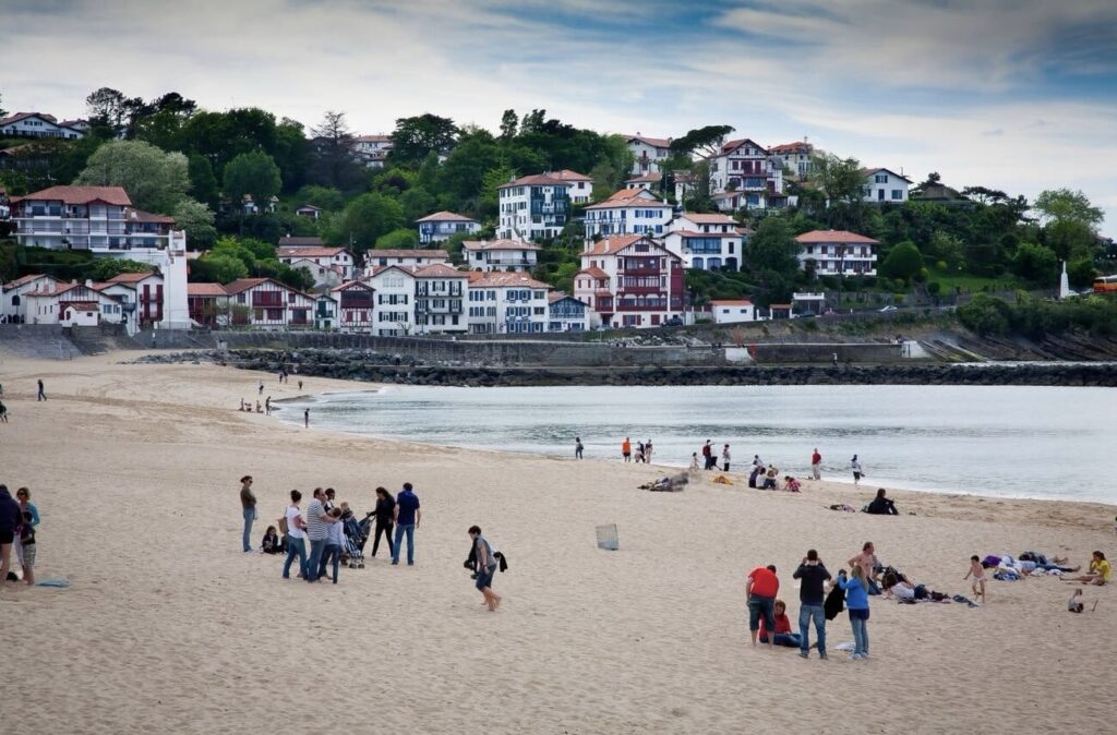 A bustling beach scene with people gathered on the sand in the foreground, with the quaint and colorful architecture of Ciboure, France, dotting the hillside in the background under a cloudy sky.