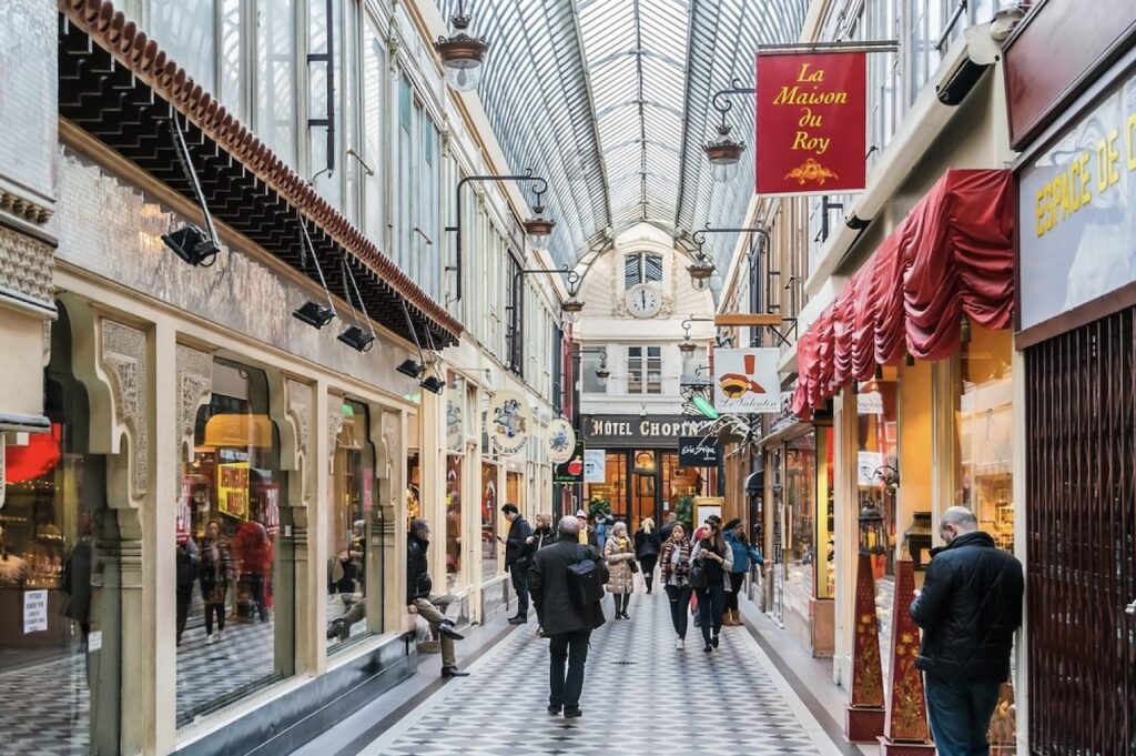 Lively interior of Passage Jouffroy in Paris, captured with shoppers walking along the black and white checkered floor. The corridor is adorned with vintage storefronts, quaint awnings, and hanging signs like 'La Maison Du Roy' adding to the historic ambiance. A clear glass roof allows natural light to highlight the detailed ironwork and timeless appeal of this 19th-century shopping arcade.