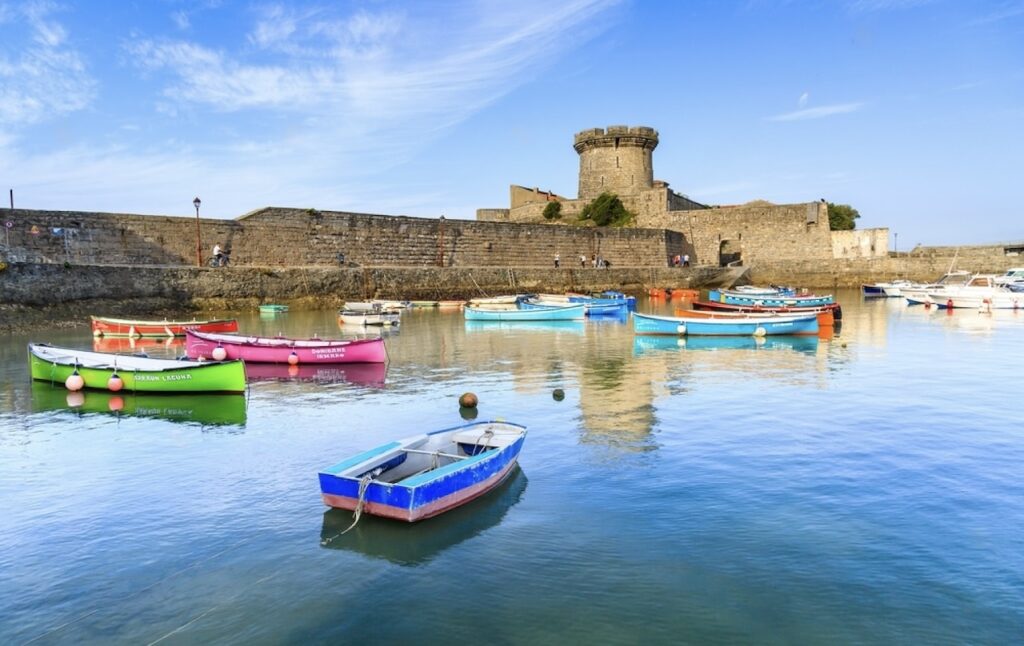 Vibrant boats moored in the serene harbor of Ciboure in the Basque Country, with the historical stone walls and round tower of the Fort of Socoa under a clear blue sky.