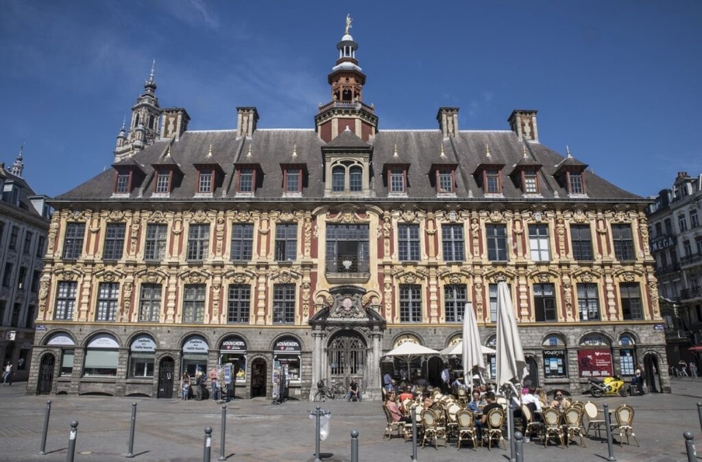 Things to Do in Lille: The Vieille Bourse, a 17th-century ornate building in Lille, France, basks in sunlight, highlighting its baroque facade as patrons enjoy a café terrace in the square.