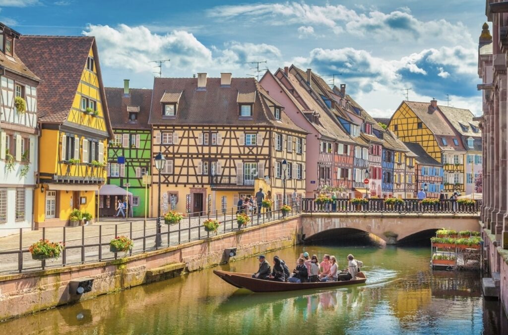 Busy pedestrian bridge over a canal in Colmar, one of the prettiest cities in France. The bridge and the canal's banks are adorned with colorful flower arrangements, enhancing the view of the charming half-timbered houses painted in pastel shades under a clear blue sky.