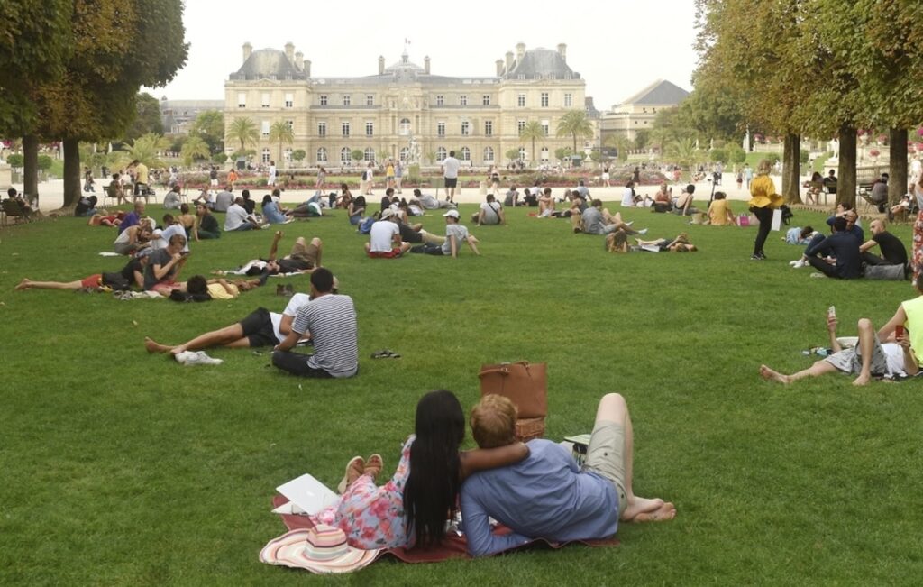 Visitors relax on the lush grass of Luxembourg Gardens in Paris, some lounging on blankets and others sitting, with the historic Luxembourg Palace in the background on a hazy day.
