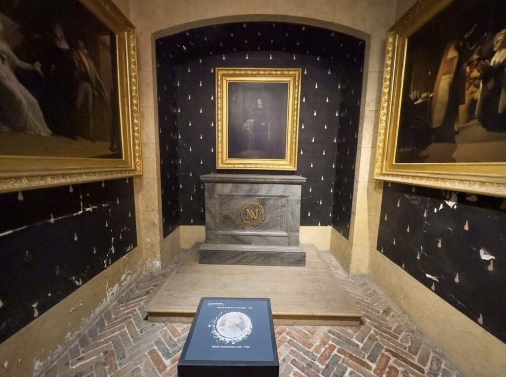 The memorial to Marie Antoinette at the Conciergerie in Paris, featuring a somber portrait placed above a stone altar with an ornate 'N' monogram. The surrounding walls are adorned with fleur-de-lis symbols on a black backdrop, with nearby gilded frames hinting at the location's royal history.