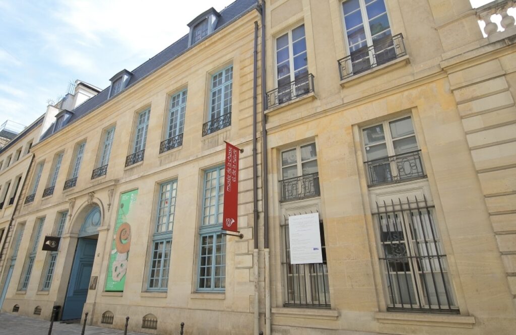hidden gems in paris: Facade of the Musée de la Chasse et de la Nature in Paris, displaying elegant limestone architecture with blue shutters, and a distinctive red banner. The museum's classic design with iron railings and large windows invites cultural exploration in the heart of the city.