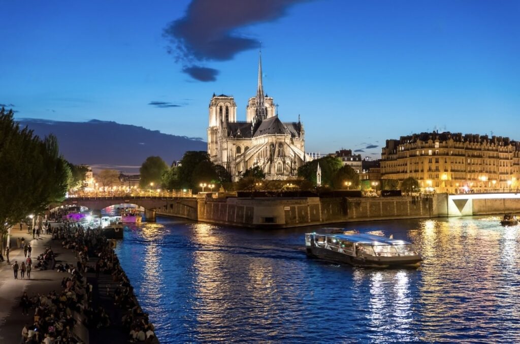Twilight descends on the Seine River in Paris, highlighting the illuminated Notre-Dame Cathedral and a cruise ship gliding by, with people gathered along the riverbank enjoying the evening ambiance.