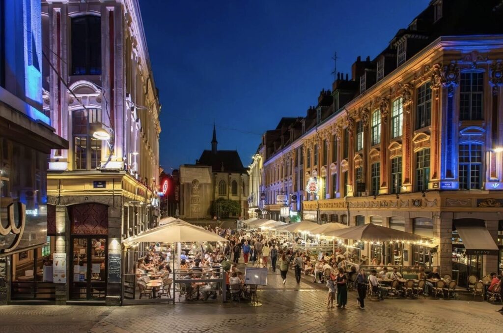 Nighttime view of a lively pedestrian street in Lille, France, with the illuminated Palais Rihour, people dining at outdoor terraces, and historic architecture under a dusk sky.