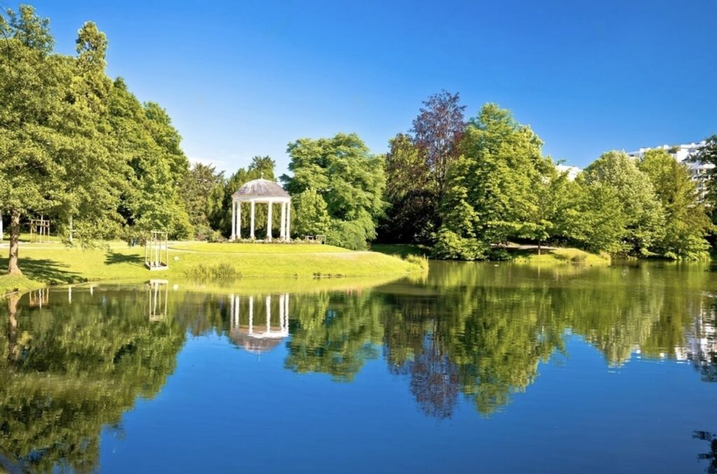 Tranquil scenery in Park de l'Orangerie, Strasbourg, showcasing a serene lake reflecting the clear blue sky and surrounding lush greenery. A classic white gazebo stands on the manicured lawn beside the water, creating a perfect symmetry with its reflection, and offers a picturesque spot for relaxation amidst nature.